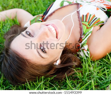 Over head portrait of an attractive young woman using technology like small headphones to listen to music while laying down on green grass in a park, smiling.