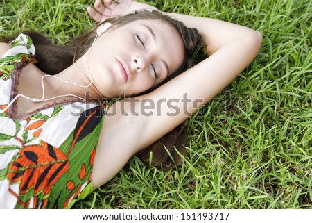 Close up portrait view of an attractive young woman laying on textured green grass listening to music with her mp3 player with her eyes closed during a summer day outdoors.