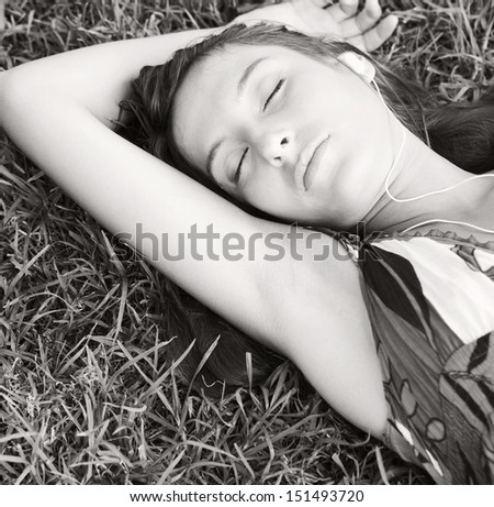 Black and white close up portrait view of an attractive young woman laying on textured green grass listening to music with her mp3 player with her eyes closed during summer.