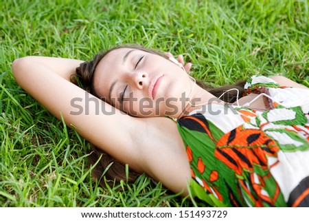 Close up portrait view of an attractive young woman laying on textured green grass listening to music with her mp3 player with her eyes closed during a summer day outdoors.