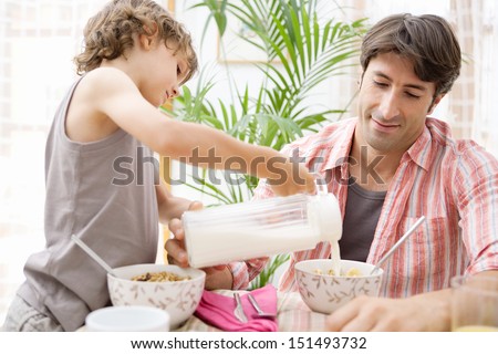 Single parent dad and his young son having breakfast together and eating cereals with the boy helping and pouring milk in his father cereal bowl during a sunny morning at home.