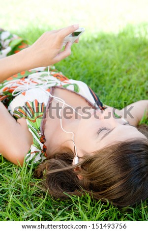 Over head close up portrait of an attractive young woman using technology electronics like an mp3 player and headphones to listen to music while laying down on green grass in a park.