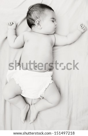 Over head full body black and white view of a baby girl wearing nappies and sleeping on a bed at home, comfortable and dreaming with her arms outstretched.