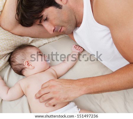 Close Up Portrait Over Head View Of A Father And His Baby Daughter Relaxing On A Bed At Home, With The Infant Sleeping And Dad Caring For Her.