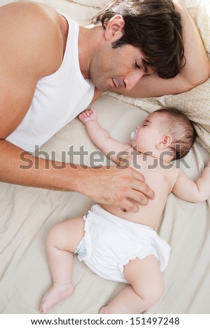 Close up portrait over head view of a father and his baby daughter relaxing on a bed at home, with the infant sleeping and dad caring for her.