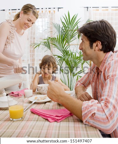 Family of three having breakfast in the morning together in their home living room, smiling and with the mother pouring milk in a glass for the son, interior.