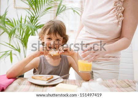 Portrait view of a mother and son together at home by a breakfast table with the boy biting into butter spread white sliced bread and mom holding a glass of orange juice, interior.