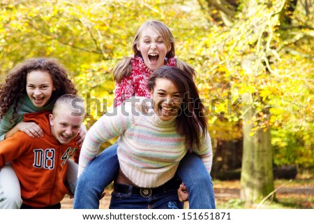 Four young teenager friends playing piggy bag racing game in a park forest during the fall autumn season with yellow trees, having fun, laughing and screaming together.