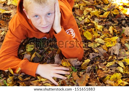 Close up portrait of a young attractive boy with freckles laying down on a bed of dry autumn tree leaves in a park forest during a sunny fall season day, outdoors.