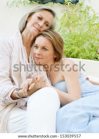 Family portrait of an adult daughter and her mature mother lounging together on a sofa in the garden at home, relaxing and hugging comfortably outdoors.