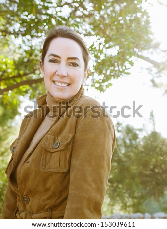 Portrait of an attractive middle aged woman visiting the forest countryside during the autumn season, wearing a leather jacket and turning to smile at camera with sun rays.