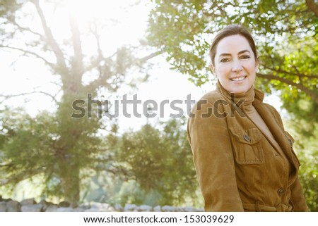 Portrait of an attractive middle aged woman visiting the forest countryside during the autumn season, wearing a leather jacket and turning to smile at camera with sun rays.