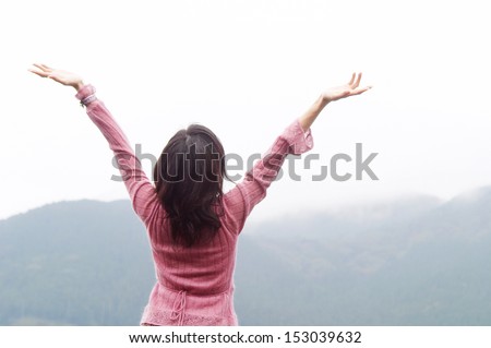 Rear view of a young asian woman standing near nature mountains with her arms stretched up in the air during a cloudy and refreshing day, breathing fresh air outdoors.