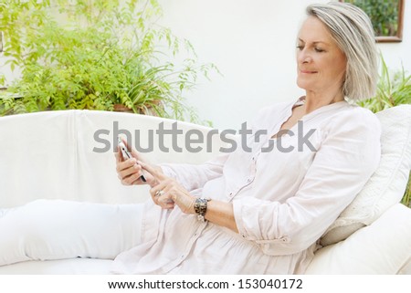 Portrait of a beautiful mature senior woman lounging on a white sofa in a garden at home and touching the screen of a smartphone device, relaxing using technology.