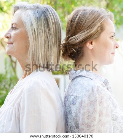 Close up family side portrait of an adult daughter with her mature mother standing back to back against each other while standing in a home garden together, outdoors.