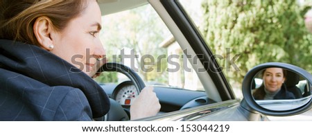 Panoramic view of an attractive mature woman sitting in a car driving and arriving at a countryside home, smiling at the camera through the rear mirror reflection on a sunny day.