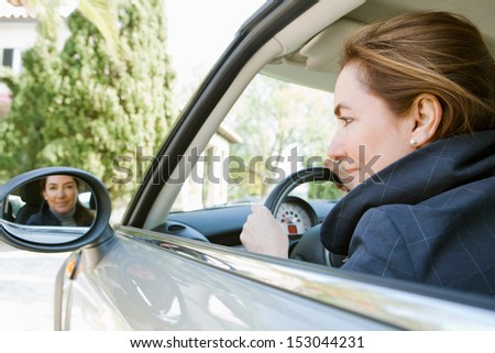 Side portrait of an attractive mature woman sitting in a car driving and arriving at a countryside home, smiling at the camera through the rear mirror reflection on a sunny day.