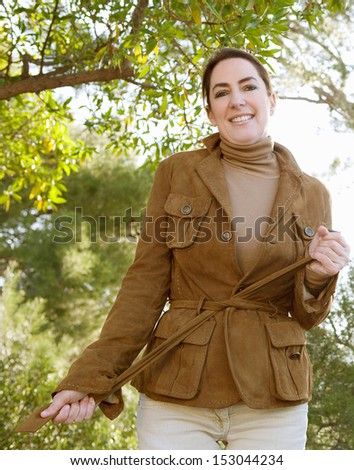 Portrait of an attractive middle aged woman visiting the forest countryside during the autumn season, wearing a leather jacket and tying the belt tight, smiling with sunshine.