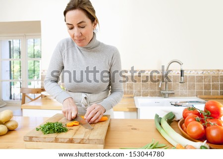 Portrait view of a middle aged woman slicing carrots with a knife on a wooden chopping board in the kitchen at home with multiple vegetables around, interior.