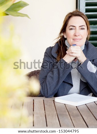 Portrait of an attractive mature woman sitting at a home garden wooden table at home, smiling at the camera during a winter day and leaning her chin on her hands, relaxing with a book.