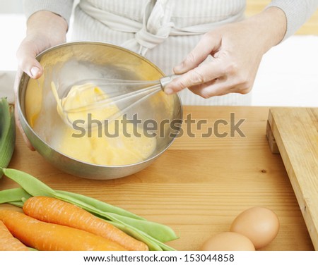 Close up detail of a woman hands beating eggs in a silver mixing bowl in the kitchen at home with multiple vegetables and eggs around, using a whisk, interior.
