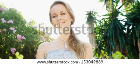 Panoramic view of a beautiful woman standing in a green garden with trees and flowers, smiling at the camera with the sun shining through her neck during a summer day.