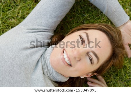 Over head close up portrait of an attractive and sophisticated mature woman wearing a grey high polo neck jumper and smiling at the camera while relaxing laying down on green grass.