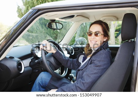 Portrait of a proud new car owner woman sitting on the driver seat with the door open, smiling at the camera and with her hand on the wheel, during a sunny cold day.