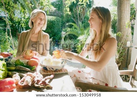 Grown up daughter and mother sitting together in a home garden peeling apples and preparing vegetables and healthy organic food during a sunny day, having a conversation.