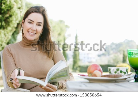 Attractive middle age woman sitting in her home garden reading a book at a breakfast table with fruit and drink during a sunny autumn day, outdoors.