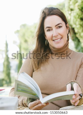Attractive and elegant middle age woman sitting in her home garden reading a book and relaxing during a sunny autumn day, outdoors.