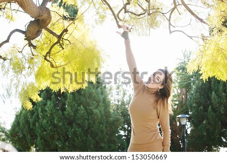 Portrait of a middle aged attractive woman wearing a thick brown jumper while visiting the countryside and reaching up a tree branch, during a sunny autumn day.