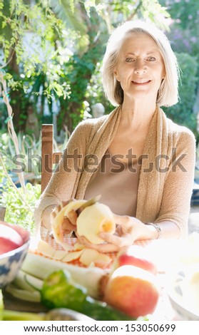 Elegant and smart mature woman sitting in a home garden preparing vegetarian food and peeling a red apple while relaxing and smiling during a sunny day, outdoors.