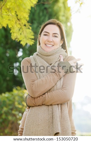 Portrait of a middle aged attractive woman wearing a thick woolly knitted brown scarf while visiting the countryside and hugging herself keeping warm, during a sunny autumn day.