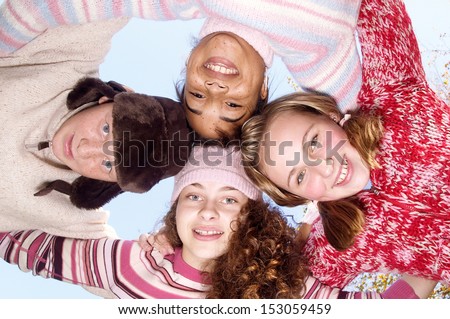Under view of four young and diverse friends with arms around each others shoulders looking and smiling at the camera together against a sunny sky, outdoors.