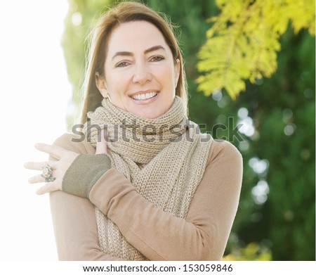 Portrait of a middle aged attractive woman wearing a thick woolly knitted brown scarf while visiting the countryside and hugging herself keeping warm, during a sunny autumn day.