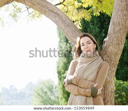 Elegant and sophisticated middle age attractive woman leaning on a tree trunk in her home garden, smiling and enjoying a sunny autumn day during the fall season, outdoors.