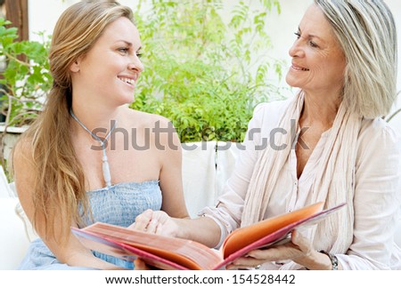 Close up portrait of a mature mother and her adult daughter flicking through a family photo album and smiling while sitting down on a white sofa in a green home garden, outdoors.