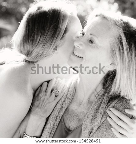 Black and white portrait of a mature mother and adult daughter spending time together in a home garden whispering and kissing during a bright and golden summer sunny day.