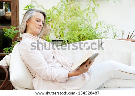Side view of an attractive mature woman lounging and relaxing at home while reading a book and sitting on a white sofa with green plants, being tranquil and serene.