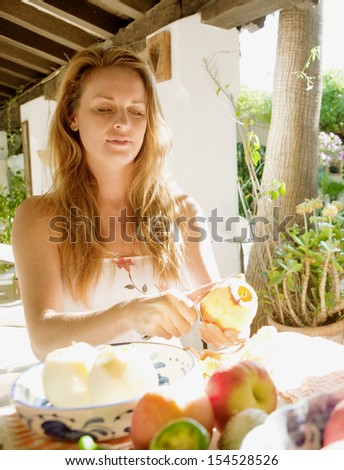 Attractive adult woman sitting at a home garden table, peeling and preparing fresh fruit and apples for cooking food during a sunny day, outdoors.