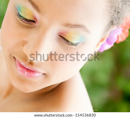 Close up beauty portrait of a young african american teenage girl wearing rainbow colors eye shadow and lipstick while standing in a green garden, turning and smiling with serenity.