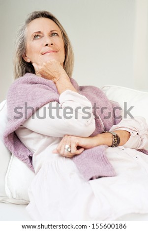 Attractive mature woman with gray hair laying and relaxing on a white sofa in a living room at home, wearing a jumper around her shoulders, keeping warm and being thoughtful.