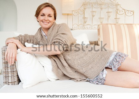 Portrait of an attractive middle aged woman laying and relaxing on a white sofa in a neutral living room at home, smiling at the camera and being serene, interior.