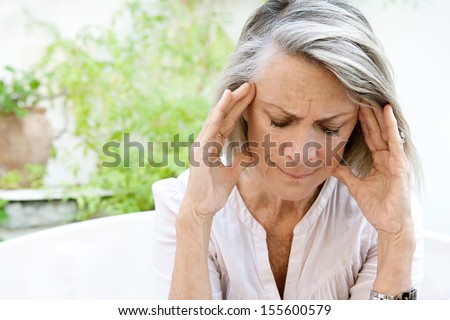 Mature Woman Sitting On A White Sofa In A Home Garden Touching Her Head With Her Hands While Having A Headache Pain And Feeling Unwell, Outdoors.