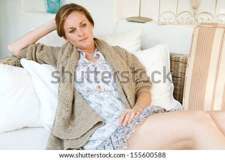 Portrait of an attractive middle aged woman laying and relaxing on a white sofa in a neutral living room at home, being thoughtful and serene, interior.