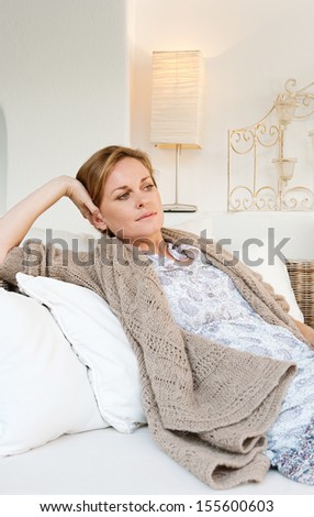 Portrait of an attractive middle aged woman laying and relaxing on a white sofa in a neutral living room at home, being thoughtful and serene, interior.