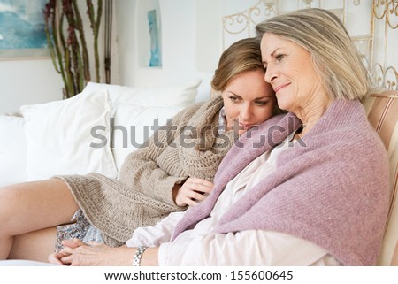 Attractive and alike adult daughter and mature mother relaxing together and being affectionate with each other while sitting on a white sofa at home and smiling, interior.