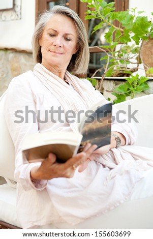 Portrait view of an attractive mature woman lounging and relaxing at home while reading a book and sitting on a white sofa with green plants, being tranquil and serene.