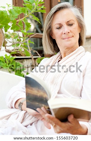 Portrait view of an attractive mature woman lounging and relaxing at home while reading a book and sitting on a white sofa with green plants, being tranquil and serene.